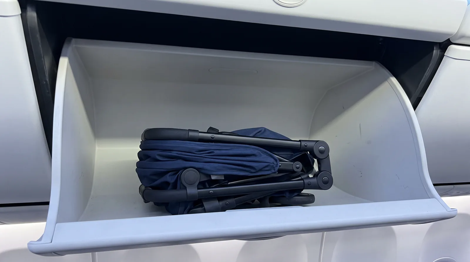A folded compact travel stroller inside an open aircraft overhead bin, with space around it.